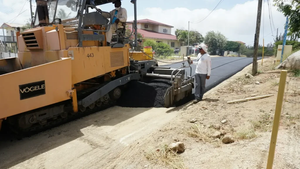 A tracked paving machine laying a strip of fresh black asphalt along a road edge while an operative checks the level