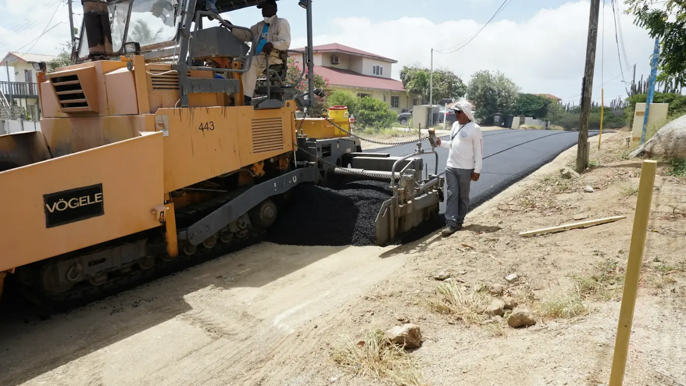 A tracked paving machine laying a strip of fresh black asphalt along a road edge while an operative checks the level