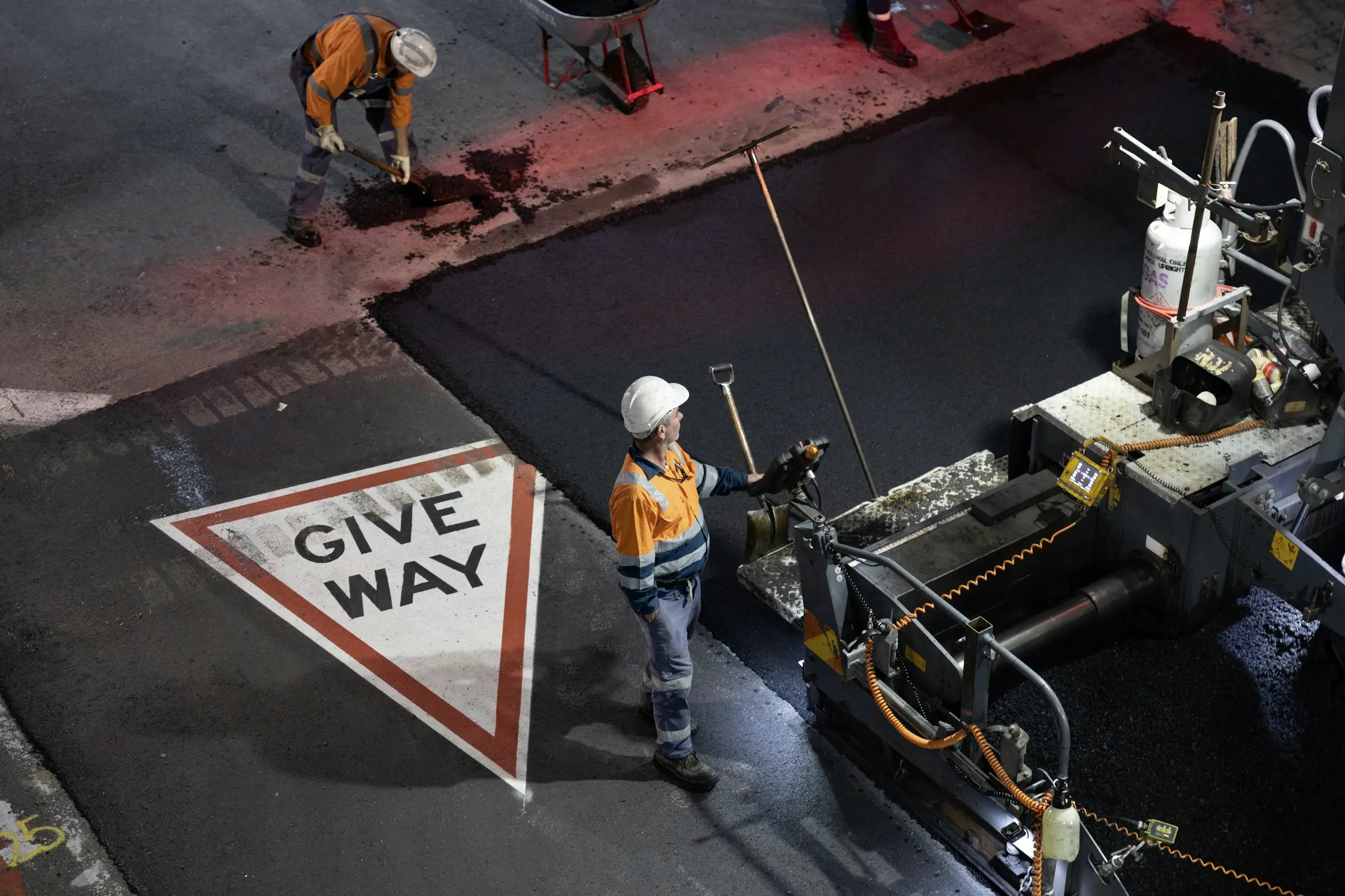 Road workers in hi-vis laying fresh asphalt behind a paving machine, with a give way marking on the old surface beside them