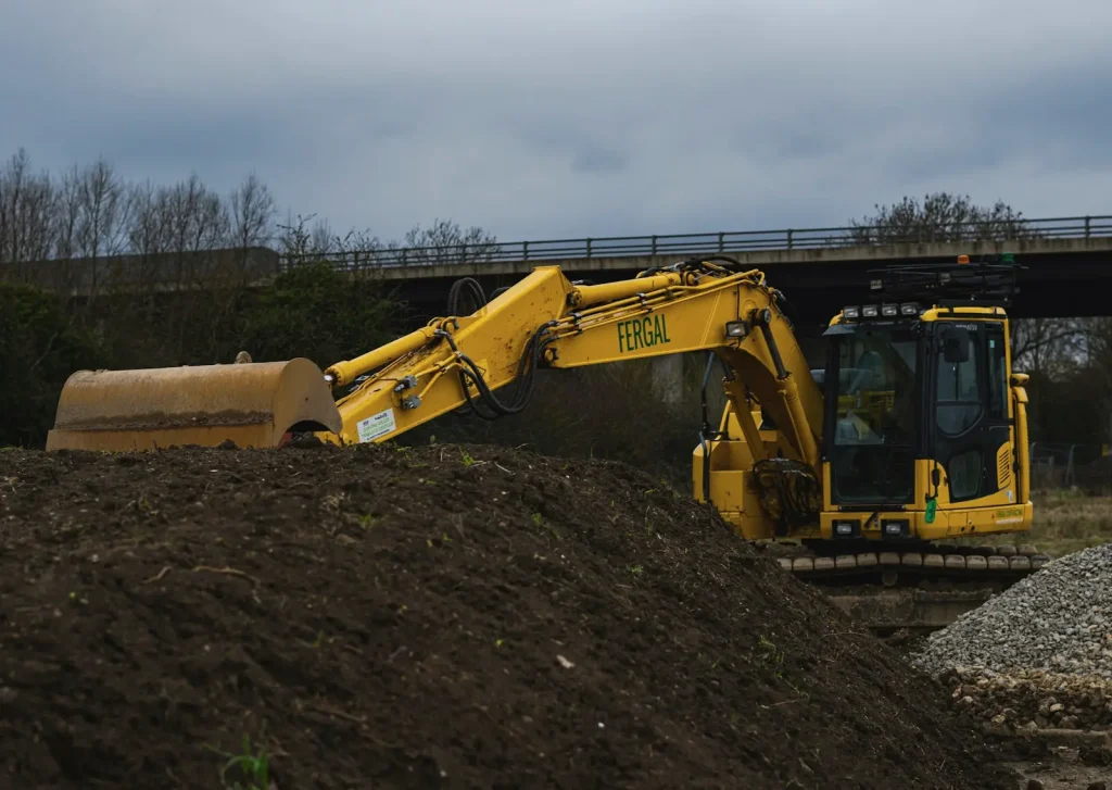 A yellow excavator resting its bucket on a stockpile of dark topsoil, with a road bridge behind