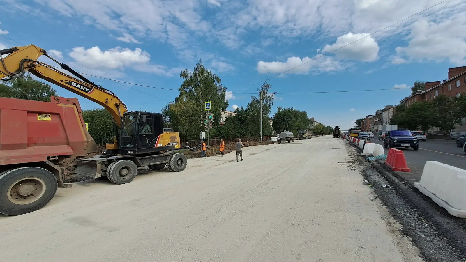 A wide road under construction at compacted sub-base level, with kerb lines set and an excavator loading a tipper lorry